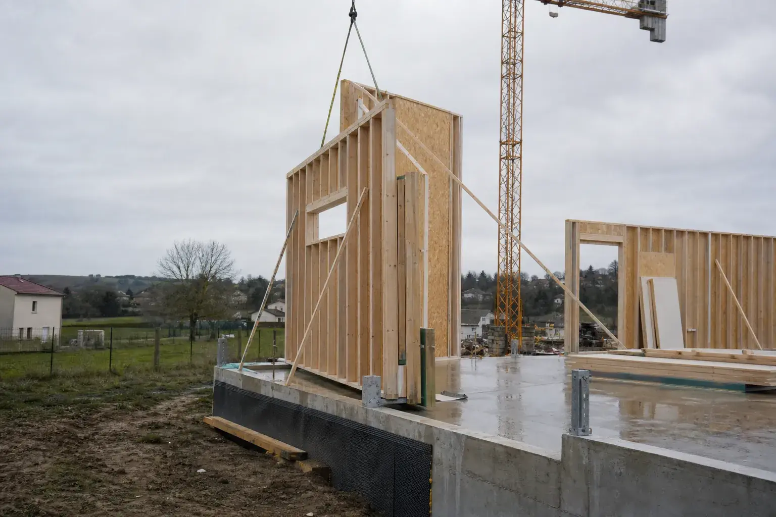 Levage d’un mur d’ossature bois sur un chantier de maison individuelle, grue au second plan