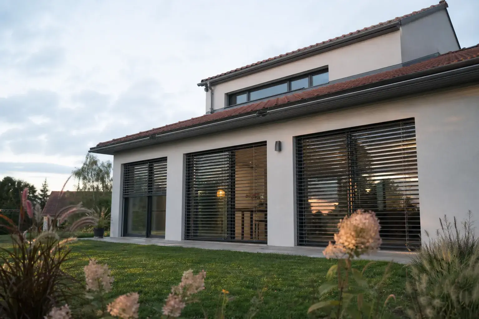 Maison individuelle neuve vue depuis le jardin en fin de journée, larges baies au sud protégées par des brise-soleil