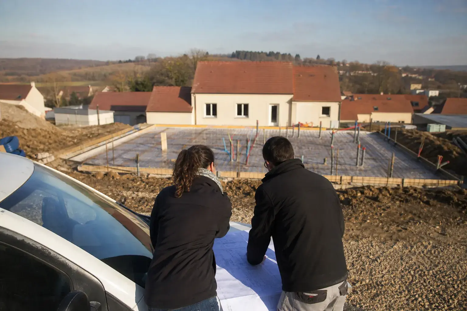 Réunion de chantier sur une dalle de maison individuelle en construction, deux personnes de dos consultant un plan sur un capot de véhicule utilitaire
