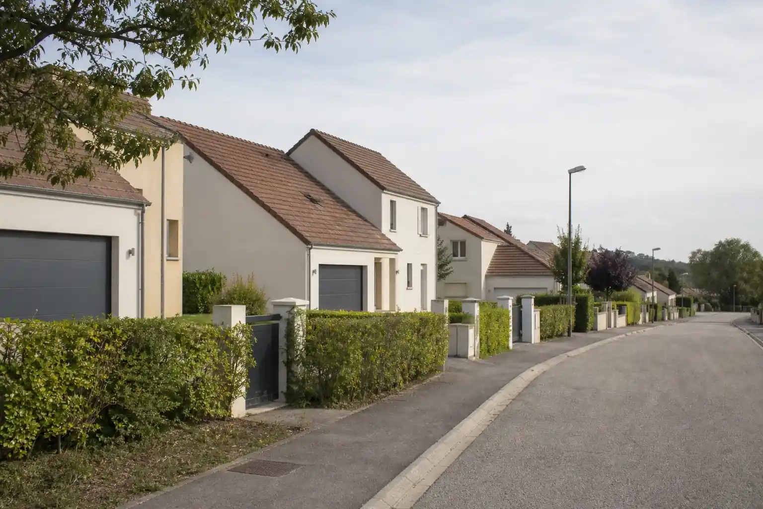 Rue résidentielle bordée de maisons individuelles récentes aux façades enduites claires, portails alignés et pelouses jeunes