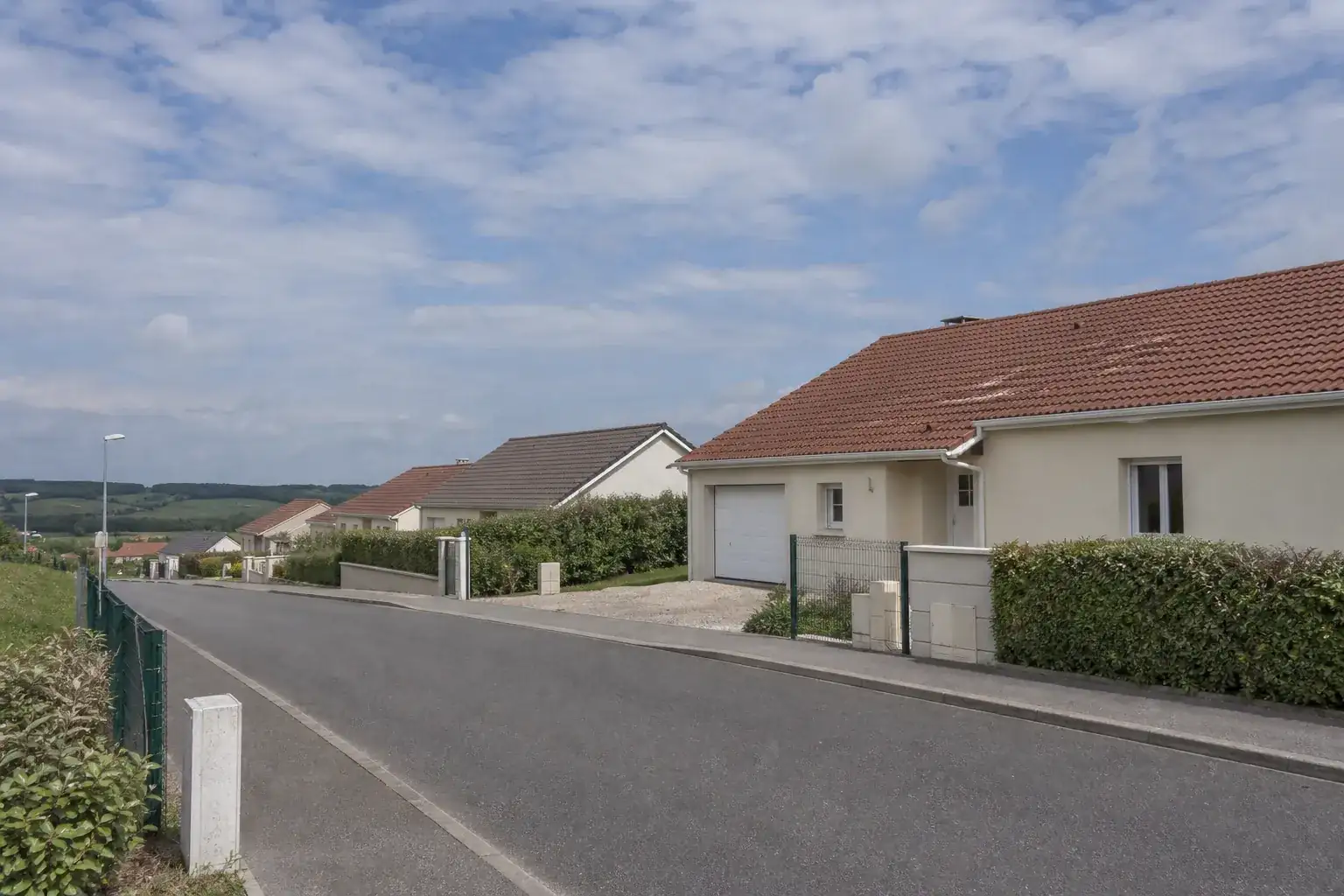 Rue résidentielle bordée de maisons individuelles récentes aux façades enduites claires, toitures en tuiles terre cuite et volets clairs