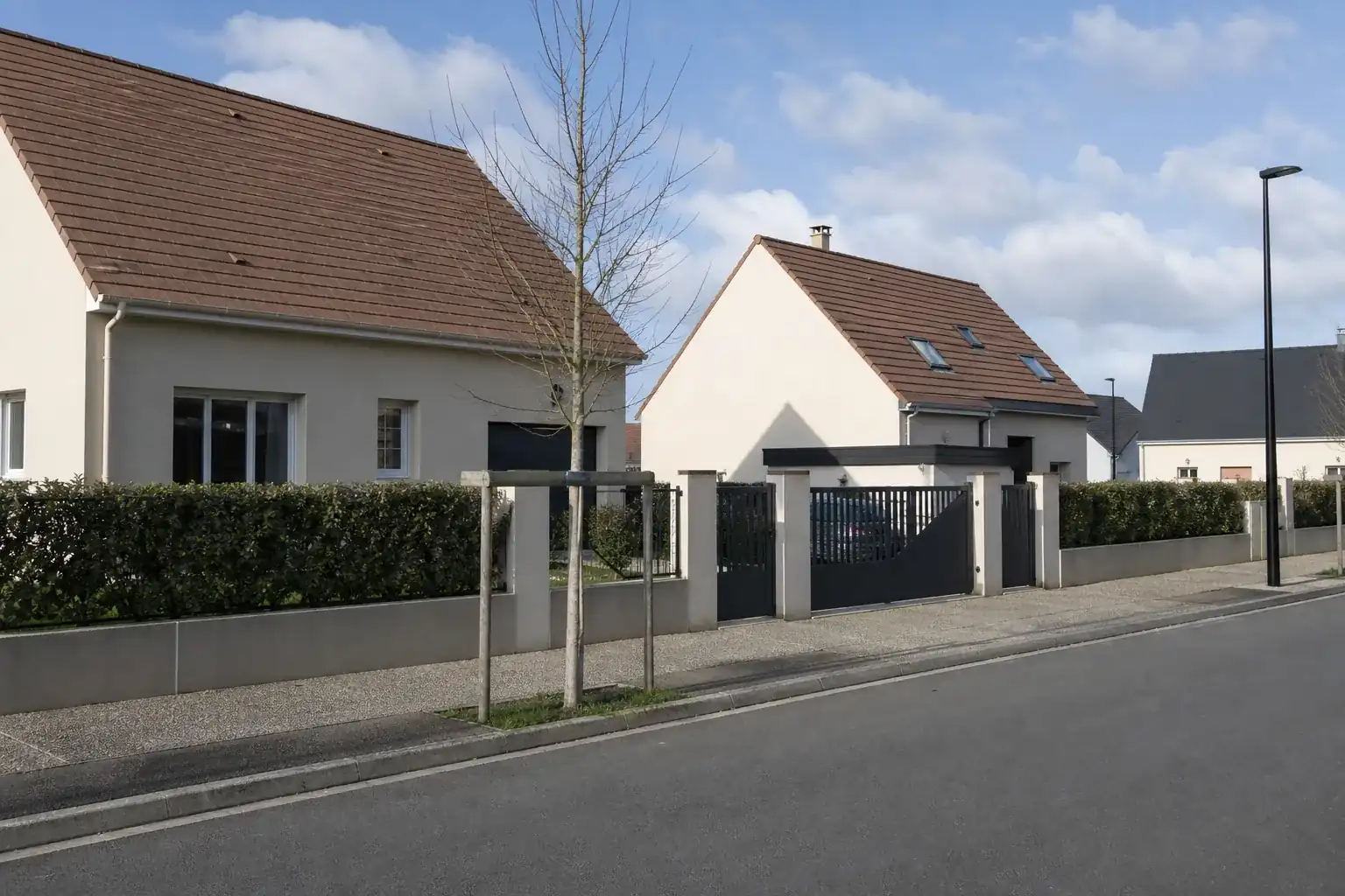 Rue résidentielle bordée de maisons individuelles récentes aux façades enduites claires, boîtes aux lettres en limite et haies fraîchement plantées